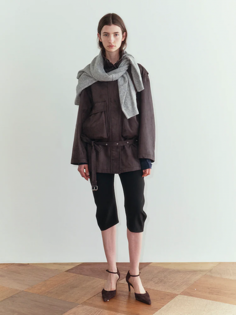 A model stands against a pale background wearing a brown suede safari jacket with large front pockets and a low-slung belt. A gray knit is draped around the shoulders, paired with black capri pants and dark brown pointed heels.