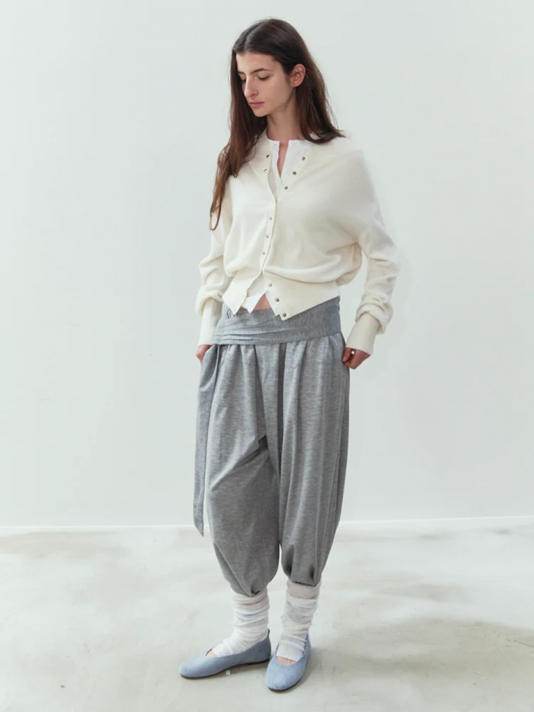 A model stands against a pale background wearing an ivory snap-button cardigan with voluminous gray pants featuring a wide folded waistband. The look is completed with layered white socks and light blue flats.