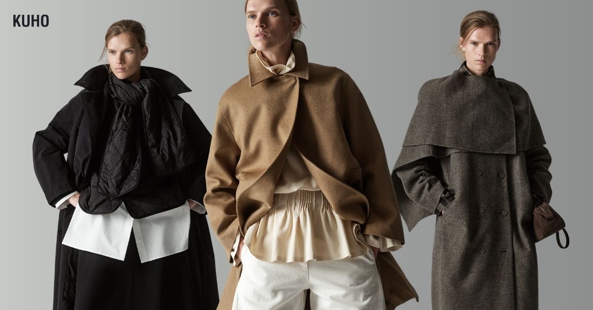 KUHO 2026 fall/winter campaign image featuring the same model in three layered looks against a light gray background. On the left, she wears voluminous black outerwear over a long white shirt and black skirt. At the center, she wears a sculptural camel jacket over a softly gathered ivory blouse and white pants. On the right, she wears a long gray double-breasted coat with a cape-like shoulder layer. The KUHO logo appears in the upper-left corner.