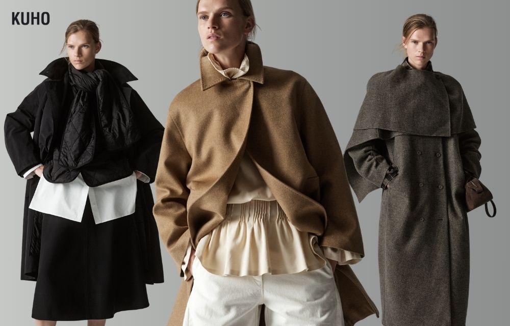 KUHO 2026 fall/winter campaign image featuring the same model in three layered looks against a light gray background. On the left, she wears voluminous black outerwear over a long white shirt and black skirt. At the center, she wears a sculptural camel jacket over a softly gathered ivory blouse and white pants. On the right, she wears a long gray double-breasted coat with a cape-like shoulder layer. The KUHO logo appears in the upper-left corner.