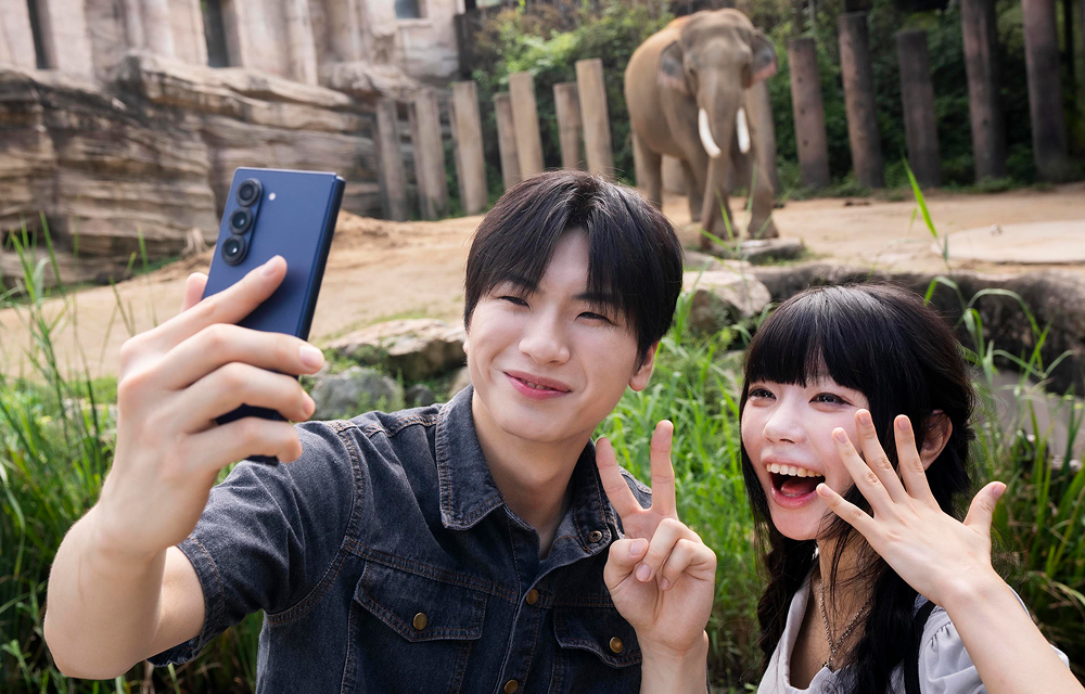 Two visitors pose for a selfie with an elephant visible in its habitat behind them at Everland’s Wild Savanna Expedition.