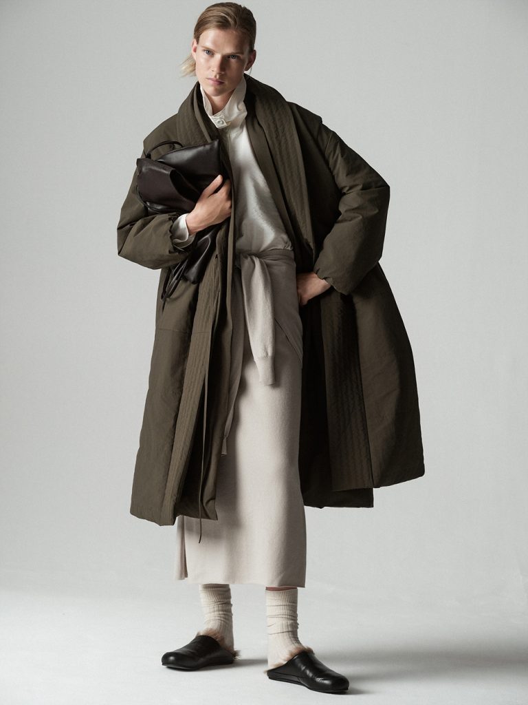 A model stands against a light gray studio background wearing an oversized dark olive long coat over an ivory high-collared top and long ivory knit dress. A light gray sweater is tied around her waist, adding another layer to the neutral-toned look. She carries a dark leather bag and wears cream socks with black slip-on shoes.