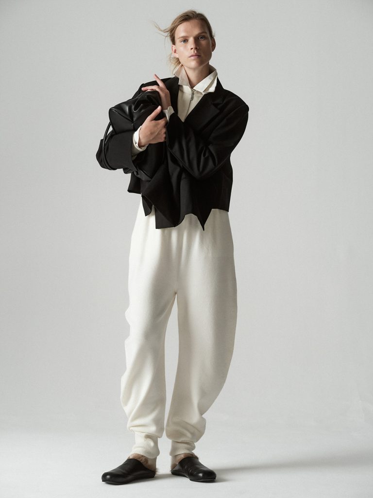A model stands against a light gray studio background wearing a cropped black jacket with an asymmetric, layered hem over an ivory high-collared shirt and voluminous ivory knit pants. She holds a large black leather bag against her chest and completes the look with black slip-on shoes.