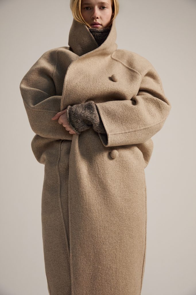 A model wears an oversized beige coat from the KUHO x Francesco Fucci 2026 winter collection, scheduled for release in November. The long coat features broad sculptural sleeves, a high overlapping collar and fabric-covered buttons. A textured dark knit is visible at the neck and cuffs against a neutral studio background.