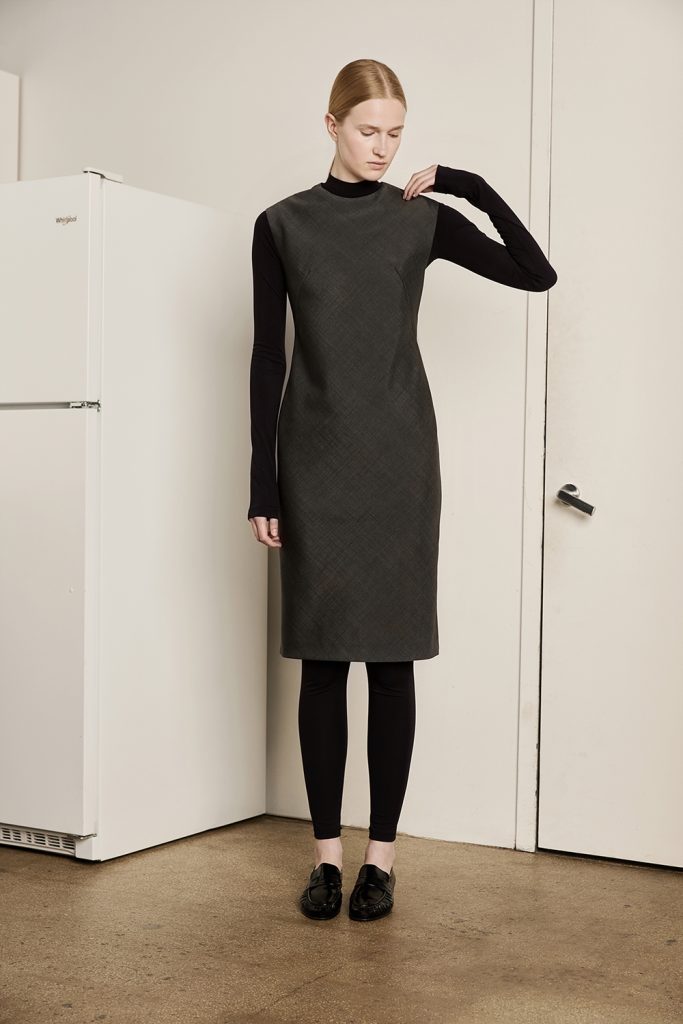 A model wears a charcoal sleeveless midi dress from the KUHO x Francesco Fucci 2026 fall collection over a fitted black high-neck top and black leggings. She completes the streamlined look with black loafers while standing between a white refrigerator and a white door in a minimalist interior.