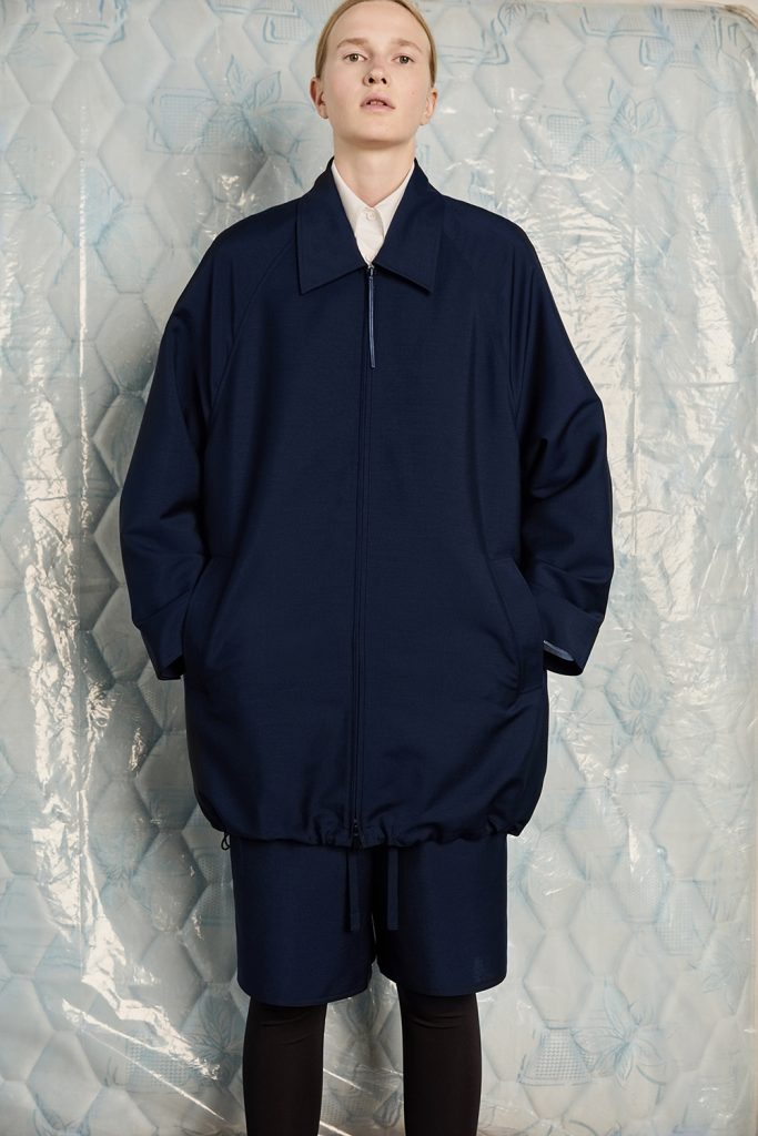 A model wears an oversized navy zip-front jacket from the KUHO x Francesco Fucci 2026 fall collection. The jacket has a broad pointed collar, rounded sleeves, large angled pockets and an adjustable drawstring hem. It is styled with matching knee-length shorts, black leggings and a white collared shirt against a pale blue quilted backdrop covered in clear plastic.