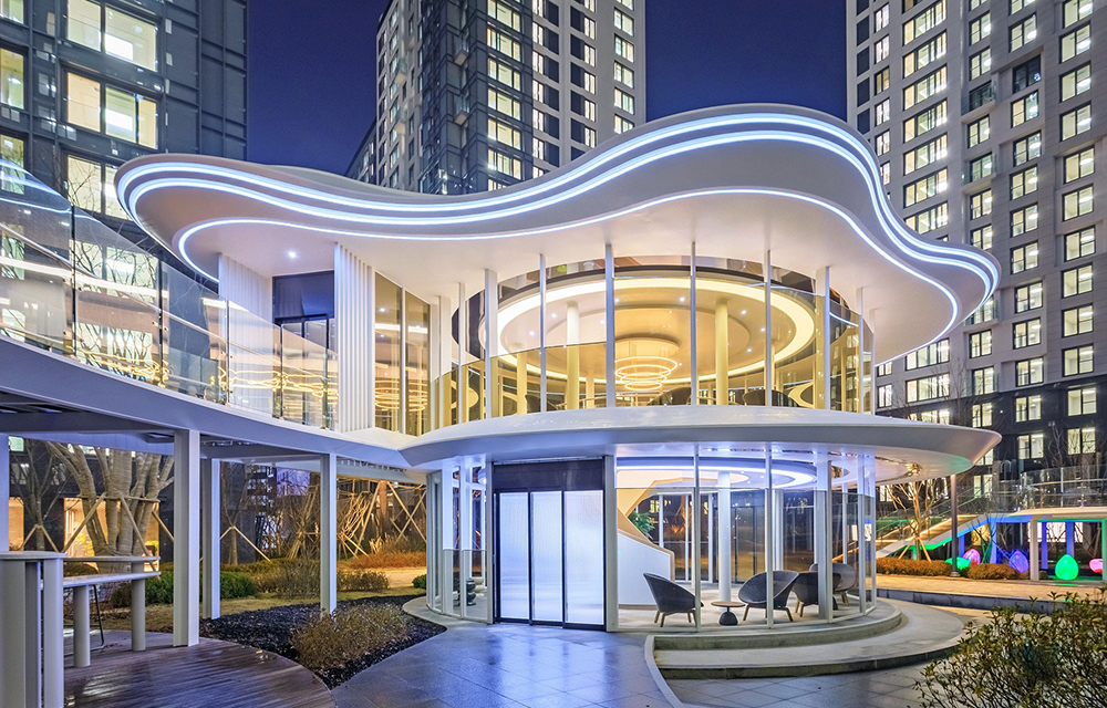 A curved, two-story glass tea house glows at night within the Blue Forest Garden, framed by high-rise residential towers and landscaped grounds.