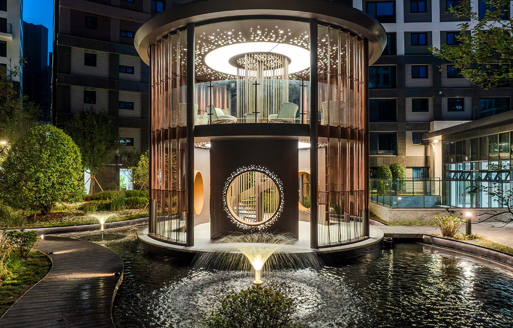 A circular illuminated pavilion rises above a water feature in the Wind Library Garden, with landscaped paths and apartment buildings surrounding the space.