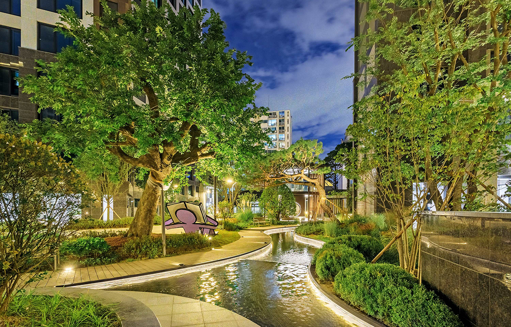 Wind Library Garden at Raemian One Perla, featuring lush landscaping, mature trees, curved walking paths, and a narrow waterway winding through the residential garden.