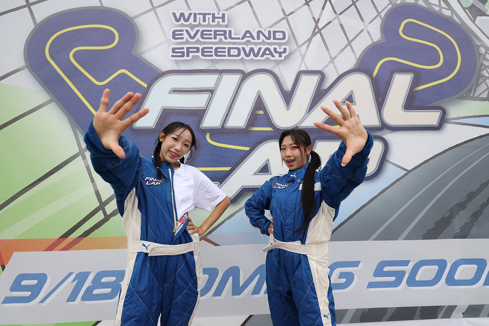 Two visitors in blue racing suits pose in front of a colorful Final Lap promotional wall at Everland