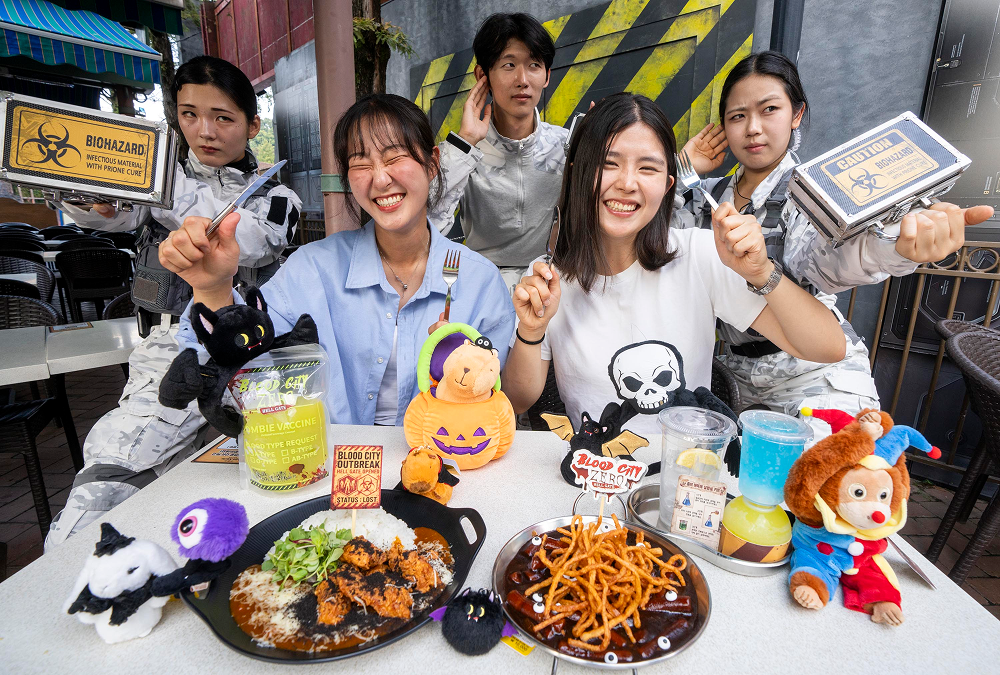 Two smiling visitors sit at an outdoor table with Blood City Zero-themed curry rice, tteokbokki, colorful drinks and Halloween character merchandise, surrounded by performers in white tactical costumes.