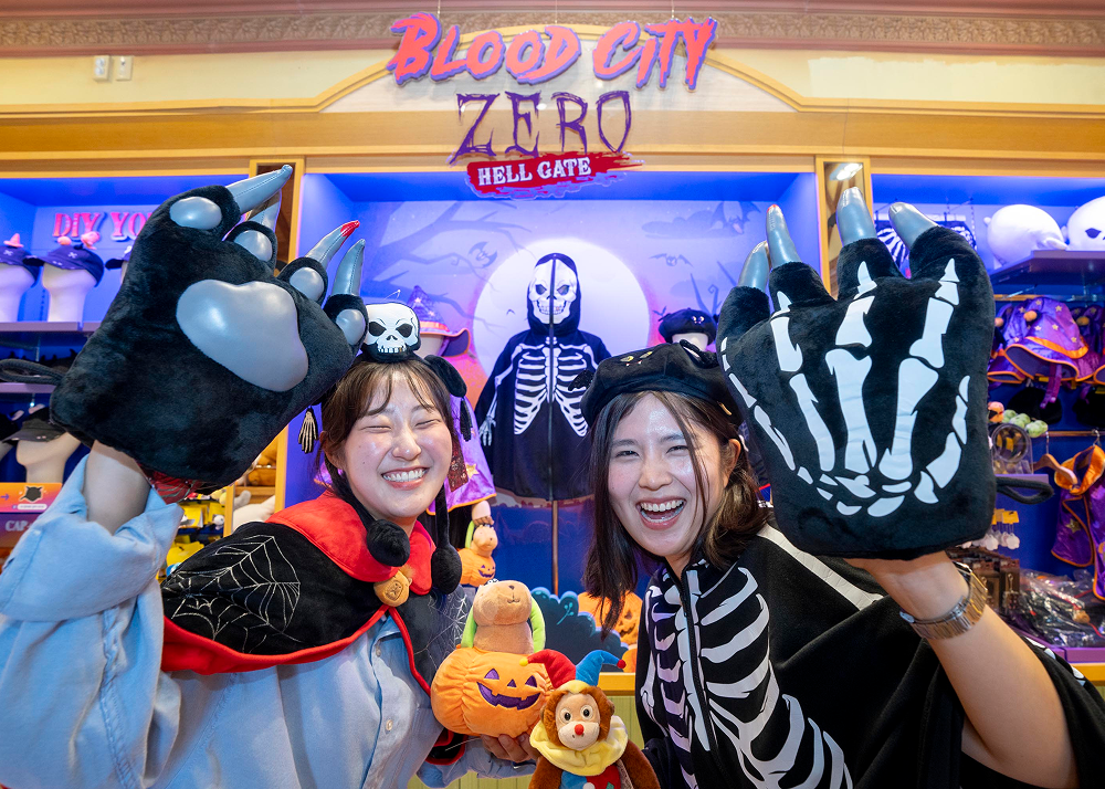 Two smiling visitors wearing skeleton-themed outfits raise oversized black skeleton gloves inside a Blood City Zero merchandise shop.