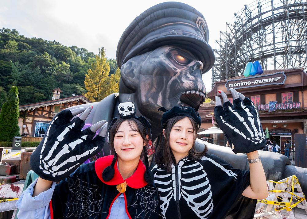 Two visitors wearing Halloween-themed outfits and oversized skeleton gloves pose in front of a giant zombie figure at Everland’s Blood City Zero.