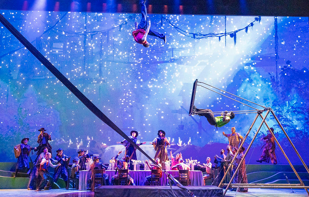 Performers take the stage beneath a starry blue backdrop as an acrobat hangs upside down above them and another swings from an aerial apparatus.