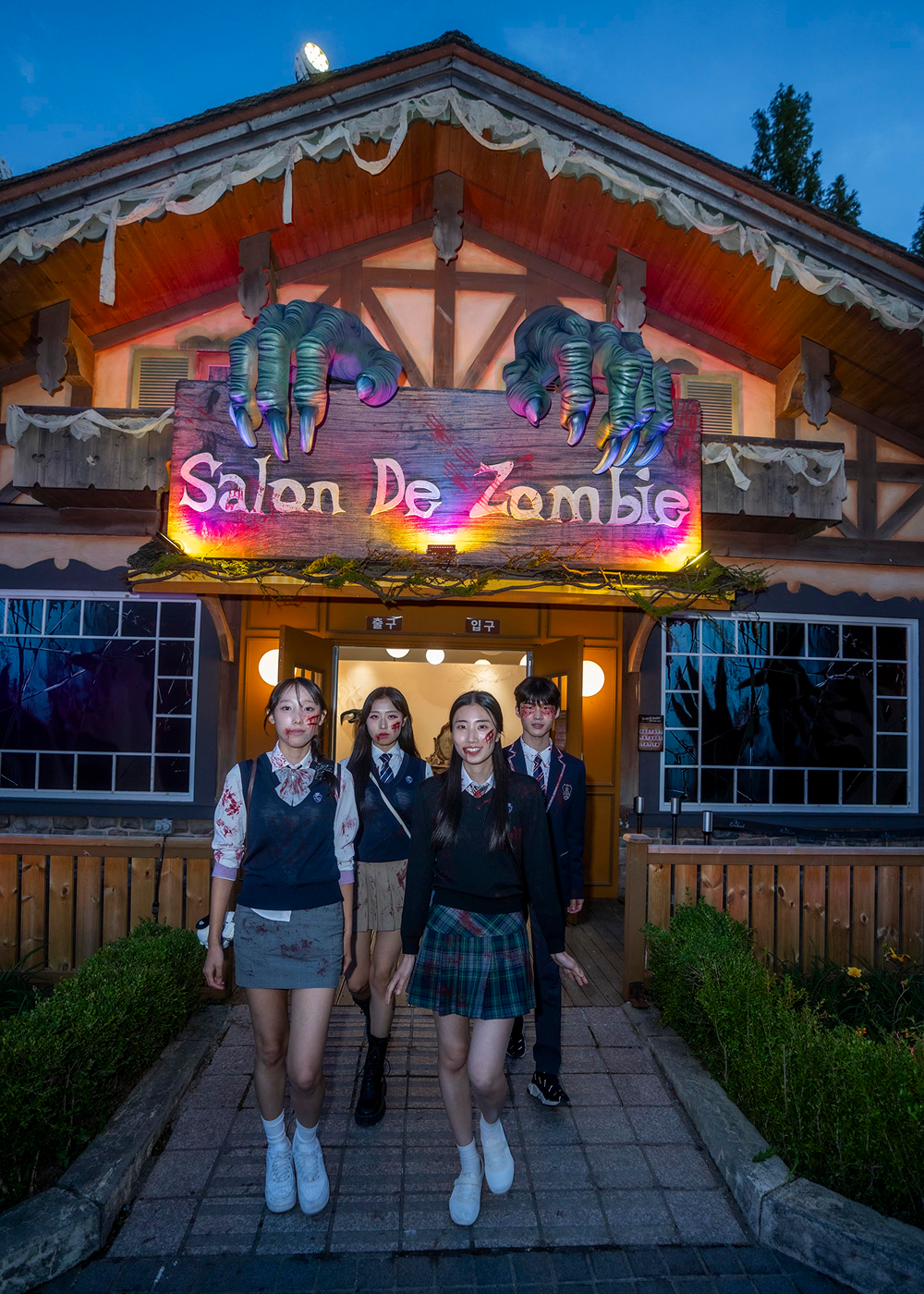 Four performers with zombie-style face makeup pose outside Everland’s Salon De Zombie beneath oversized monster hands and an illuminated sign