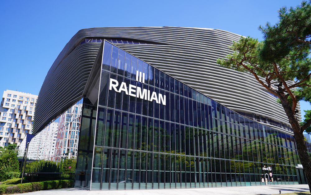 Raemian’s new black-and-white logo, featuring three vertical lines above the RAEMIAN wordmark, displayed on the glass façade of Raemian Gallery under a clear blue sky