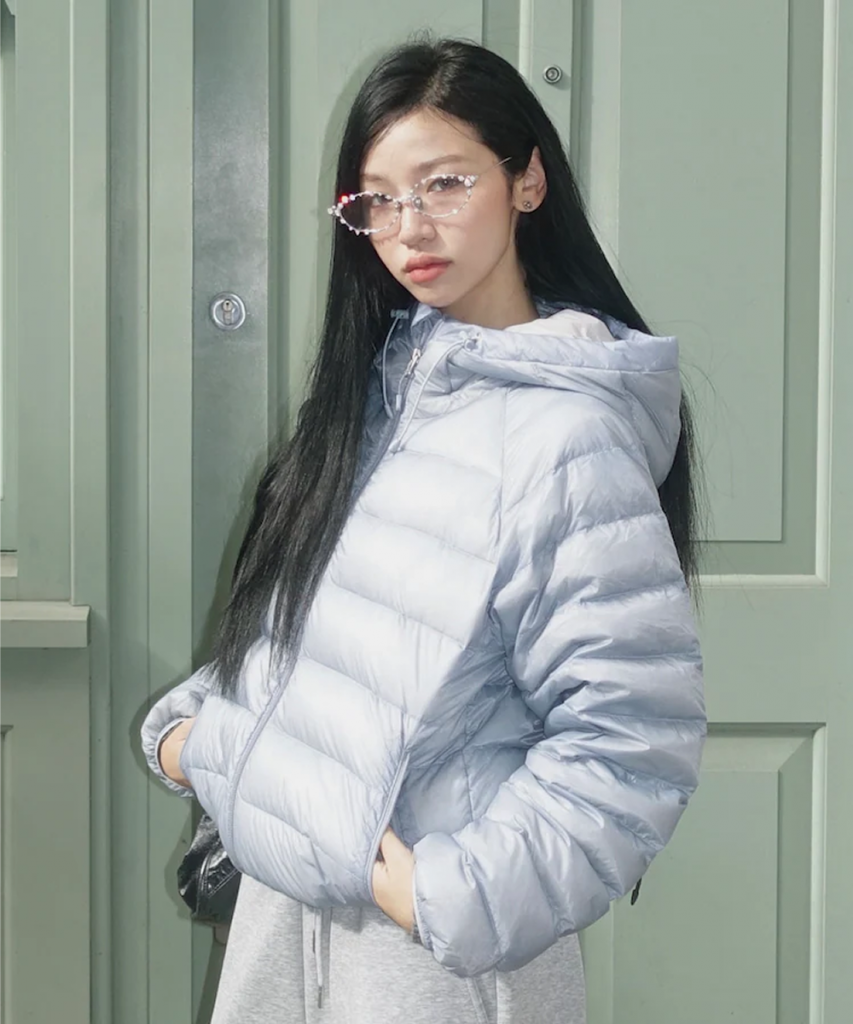 A model wears a pale blue hooded puffer jacket with gray sweatpants while standing in front of a muted green exterior wall.