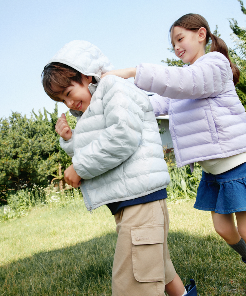 A girl in a pale lavender puffer jacket playfully adjusts the hood of a boy wearing a light blue puffer jacket outdoors.