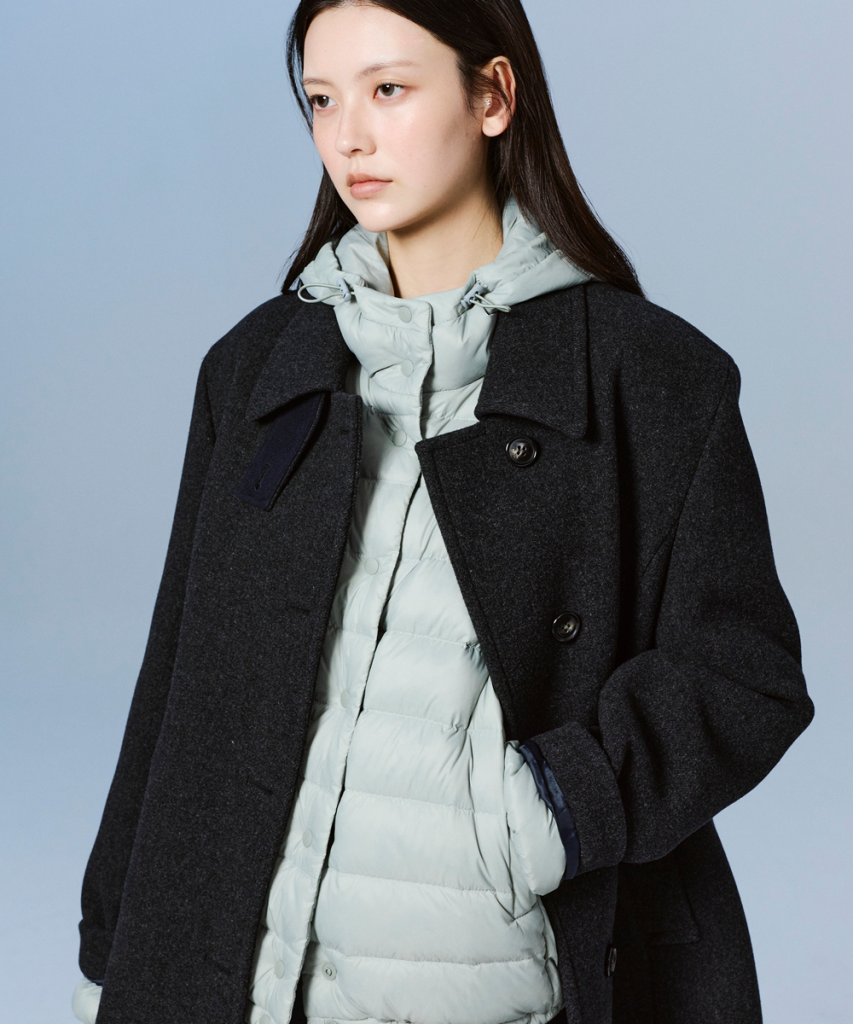 A model wears a pale blue lightweight puffer layered under a dark charcoal coat against a light blue studio background. 
