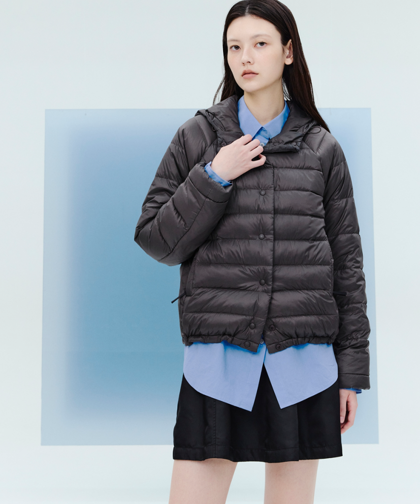 A model wears a charcoal hooded puffer jacket over a bright blue shirt and black shorts against a white and pale blue studio background.