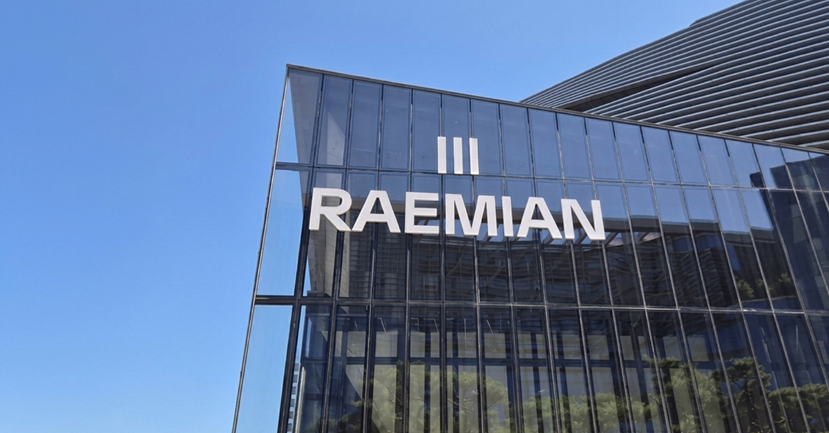 The white RAEMIAN wordmark and three-line symbol are mounted on the glass façade of a modern building against a clear blue sky.