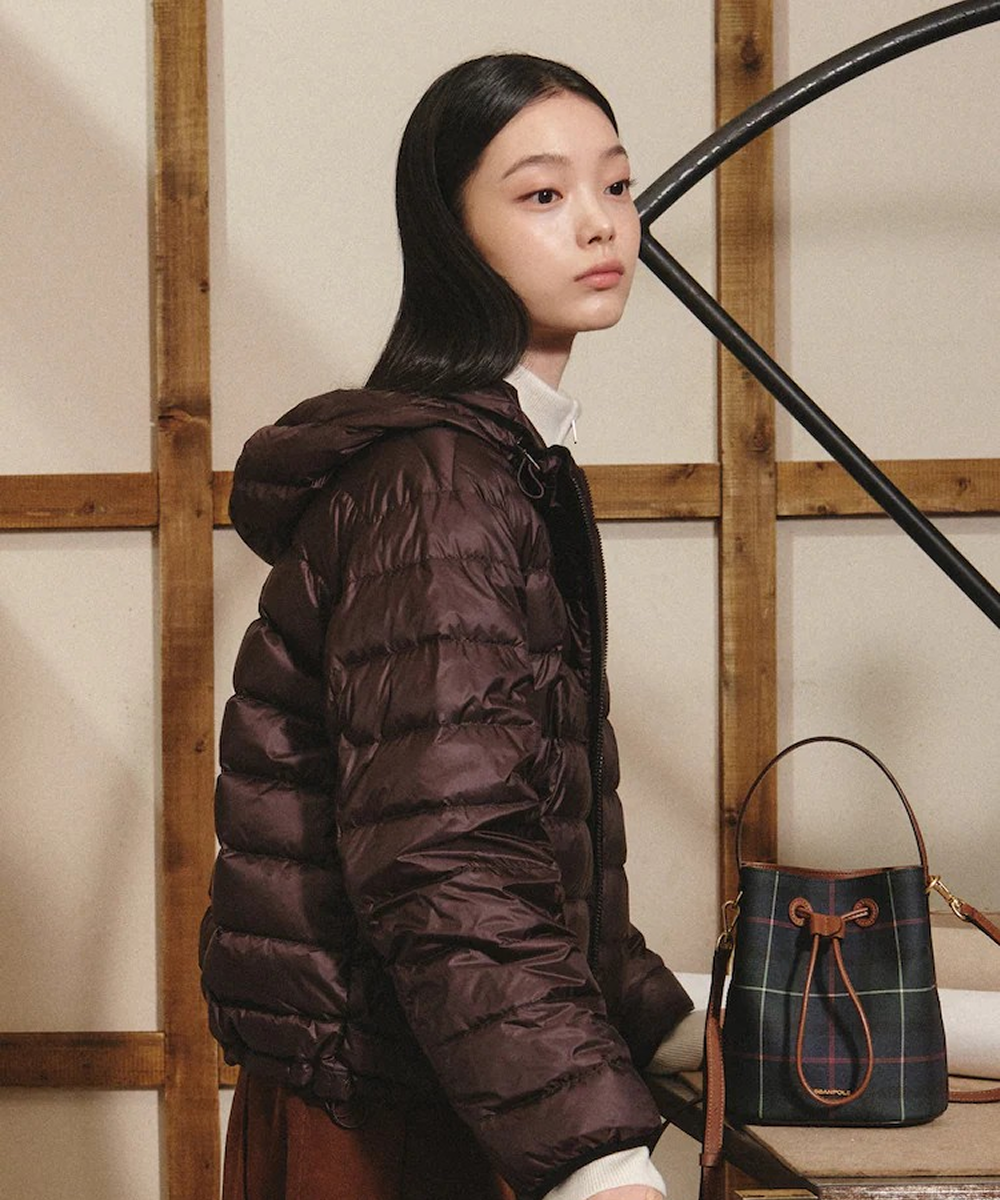 A model wears a dark brown hooded puffer jacket over an ivory high-neck top and brown wide-leg pants, carrying a small check-patterned bag. 